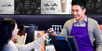 A woman paying at the counter of a coffee shop with digital signage, meant to represent the advantages of digital signage vs print signage