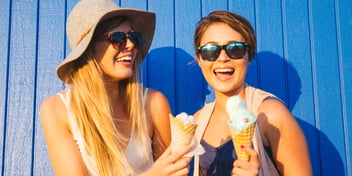 Two smiling women wearing sunglasses and eating ice cream in front of a blue paneled wall