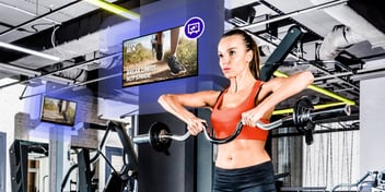 A woman lifting weights in a gym in front of screens with DOOH advertising, meant to represent the power of commerce media and in-location retail media